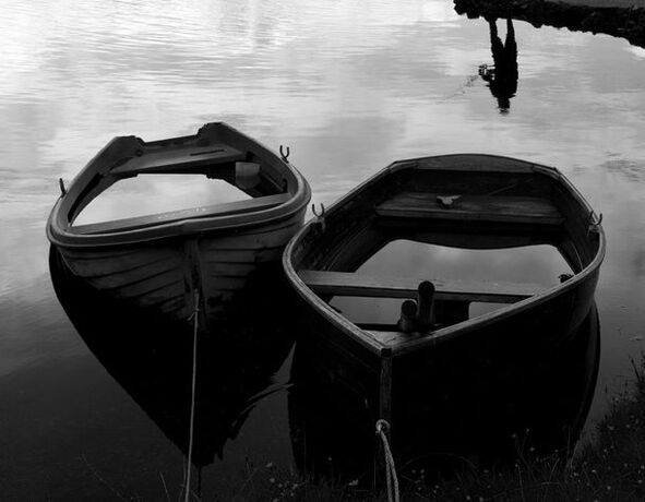 An upside down reflection of a man fishing with two waterfilled boats in the foreground