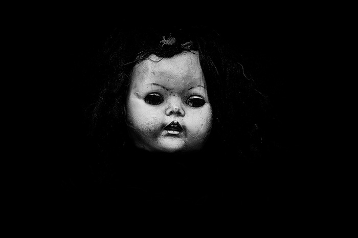 A decrepid vintage doll with a miserable looking face stares out from black negative space