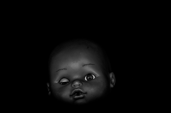 Doll face with one eye closed and one open, surrounded by black negative space