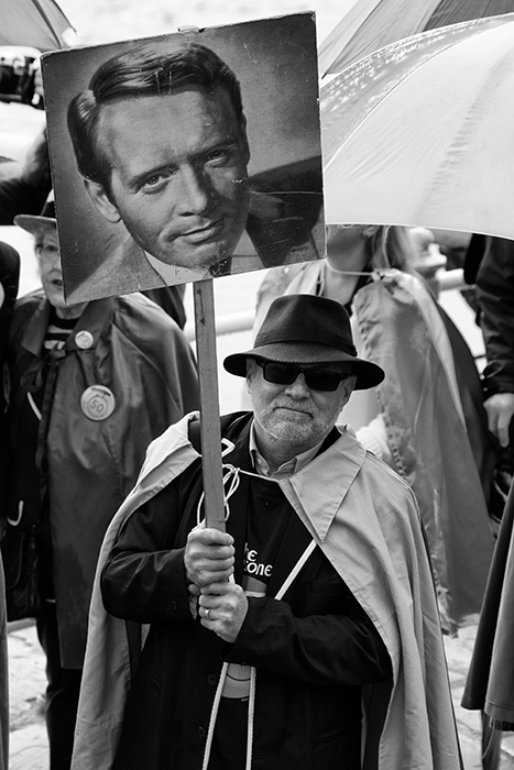 Man in cape and hat holds up a placard with the picture of No.6