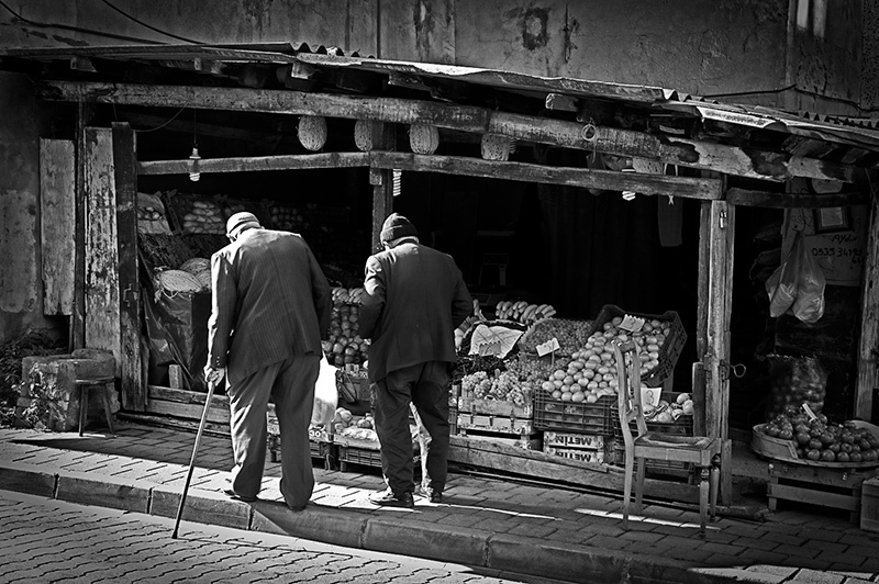 Two elderly men walk past a ramshackle fruit and veg store in strong sunlight in an Istanbul suburb