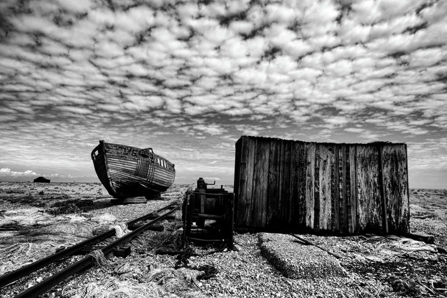A decaying fishing boat, winch and wooden hut sit under a mackerel sky on a shingle beach at Dungeness