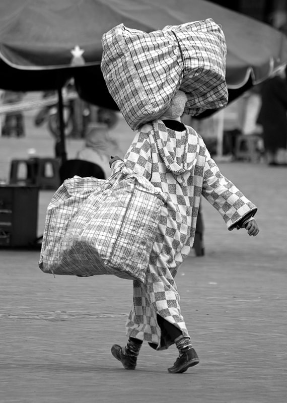 Woman in chequered North African clothing carries chequered bags, one of which is balanced on her head