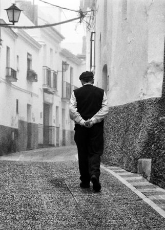 An elderly man walks away with his arms behind his back down a traditional Spanish village street